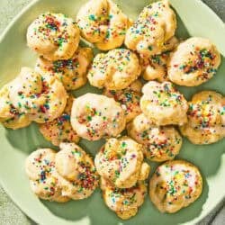 Italian lemon cookies on a platter next to a bowl of non-pareils, a stack of 2 plates, and a lemon.