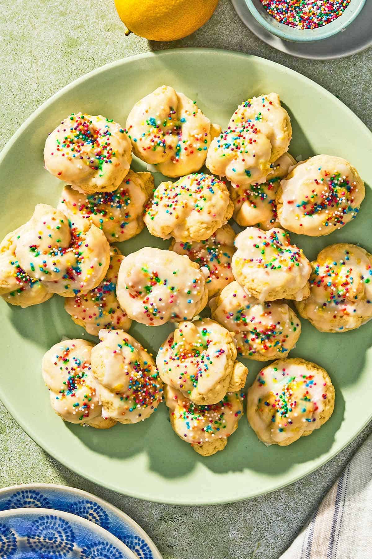 Italian lemon cookies on a platter next to a bowl of non-pareils, a stack of 2 plates, and a lemon.