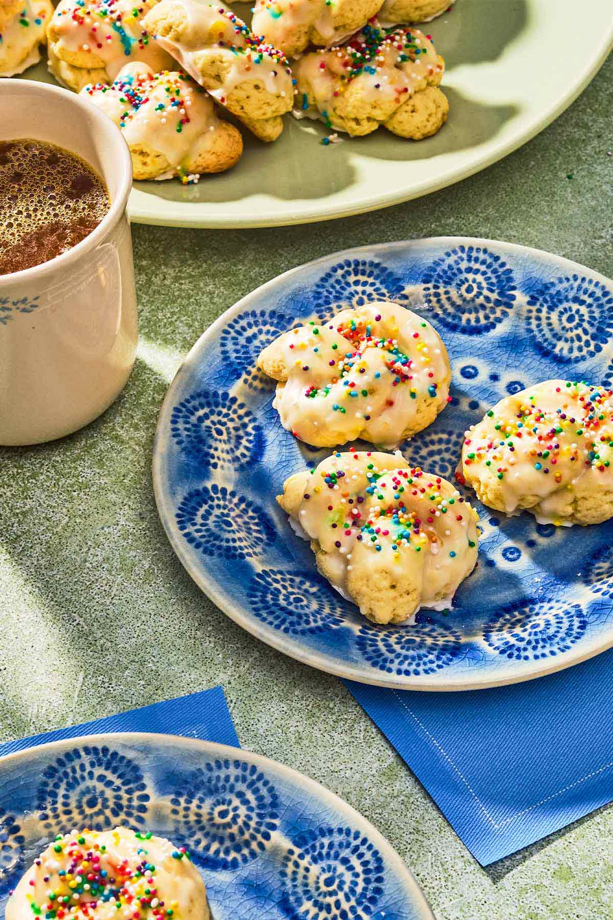 Three Italian lemon cookies on a plate, next to another plate with a cookie, a platter with the rest of the cookies, and a cup of coffee.