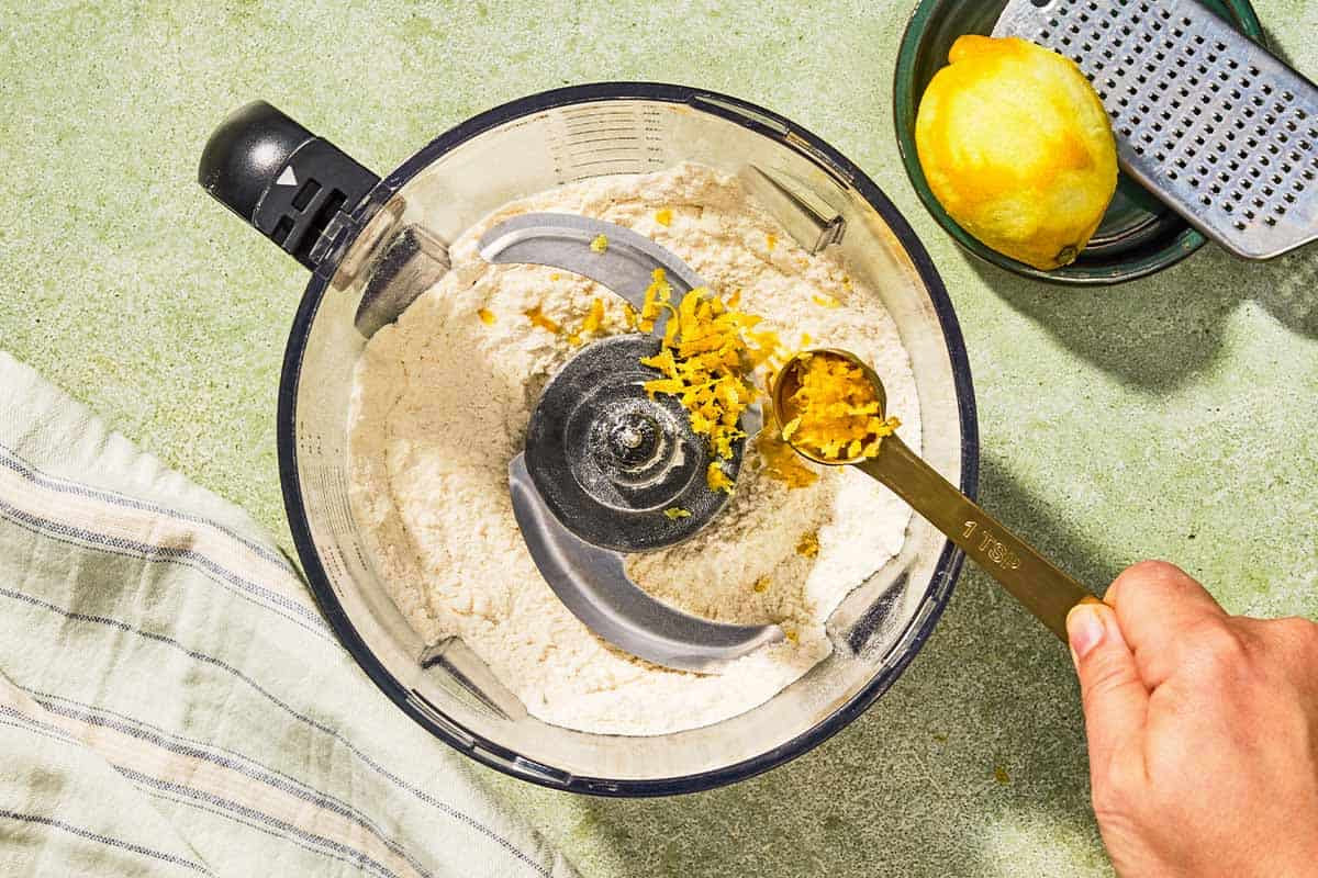 Lemon zest being added to the dry ingredients for the Italian lemon cookies in the bowl of a food processor fitted with a blade. Next to this is a kitchen towel, a zested lemon and a zester.