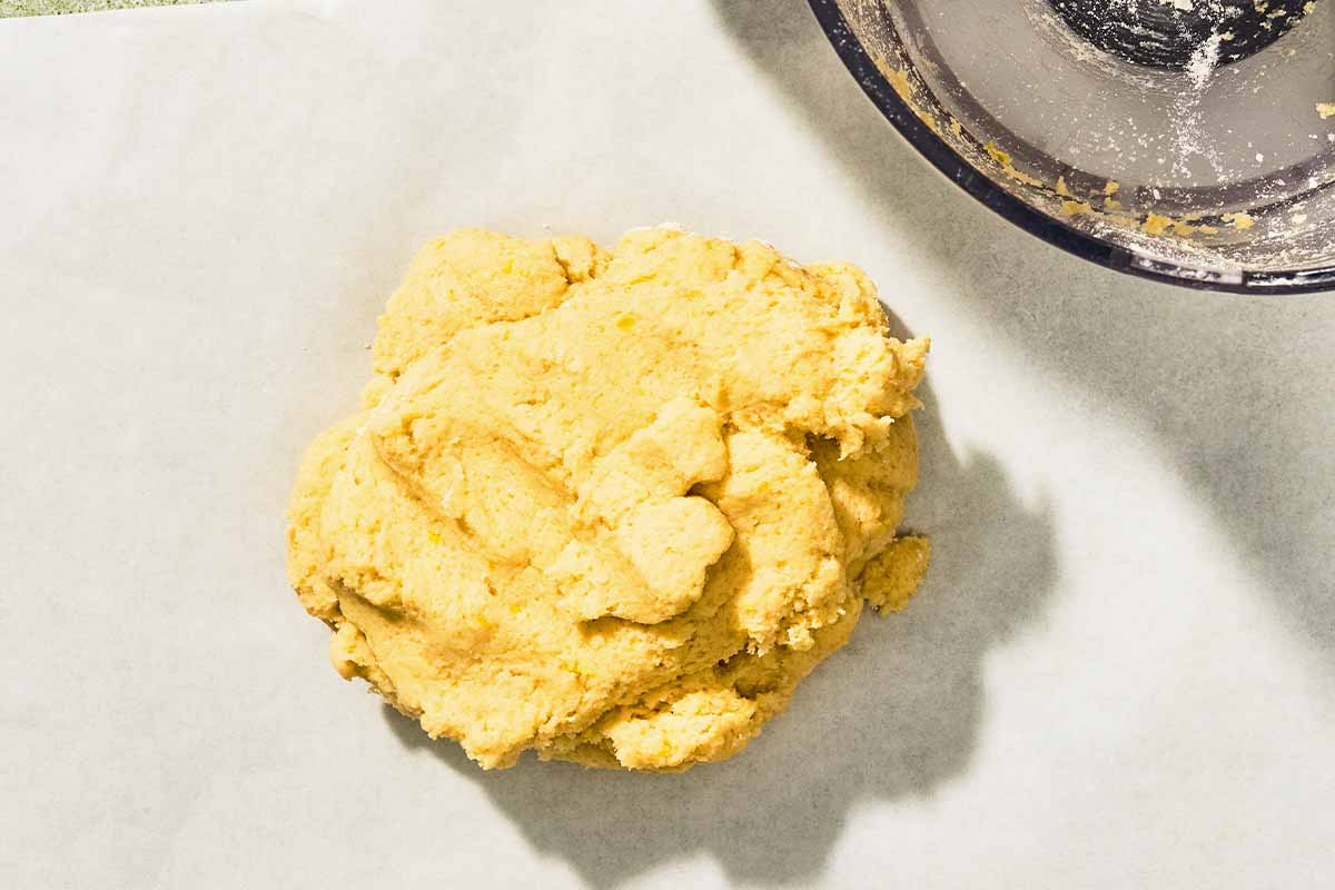 The dough for the Italian lemon cookies on a piece of parchment paper next to the bowl of a food processor.