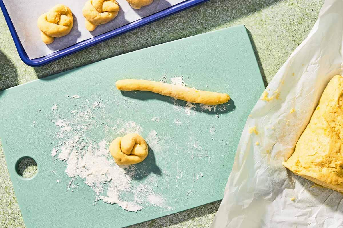 One piece of the dough for the Italian lemon cookies rolled into a log, and one shaped into a knot on a cutting board. Next to this is the rest of the dough on a piece of parchment paper and some knotted pieces of dough on a parchment lined sheet pan.