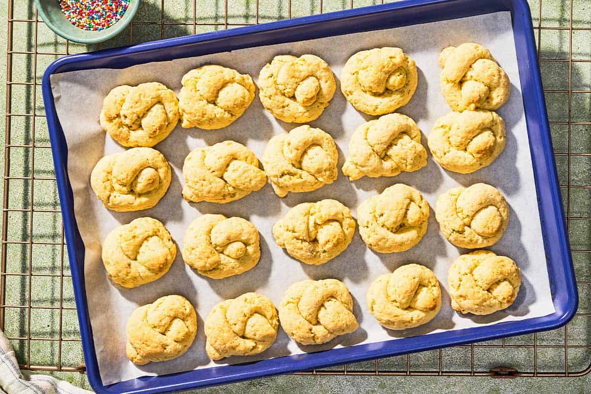 Baked, undecorated Italian lemon cookies on a parchment lined sheet pan resting on a wire rack with a bowl of non-pareils.