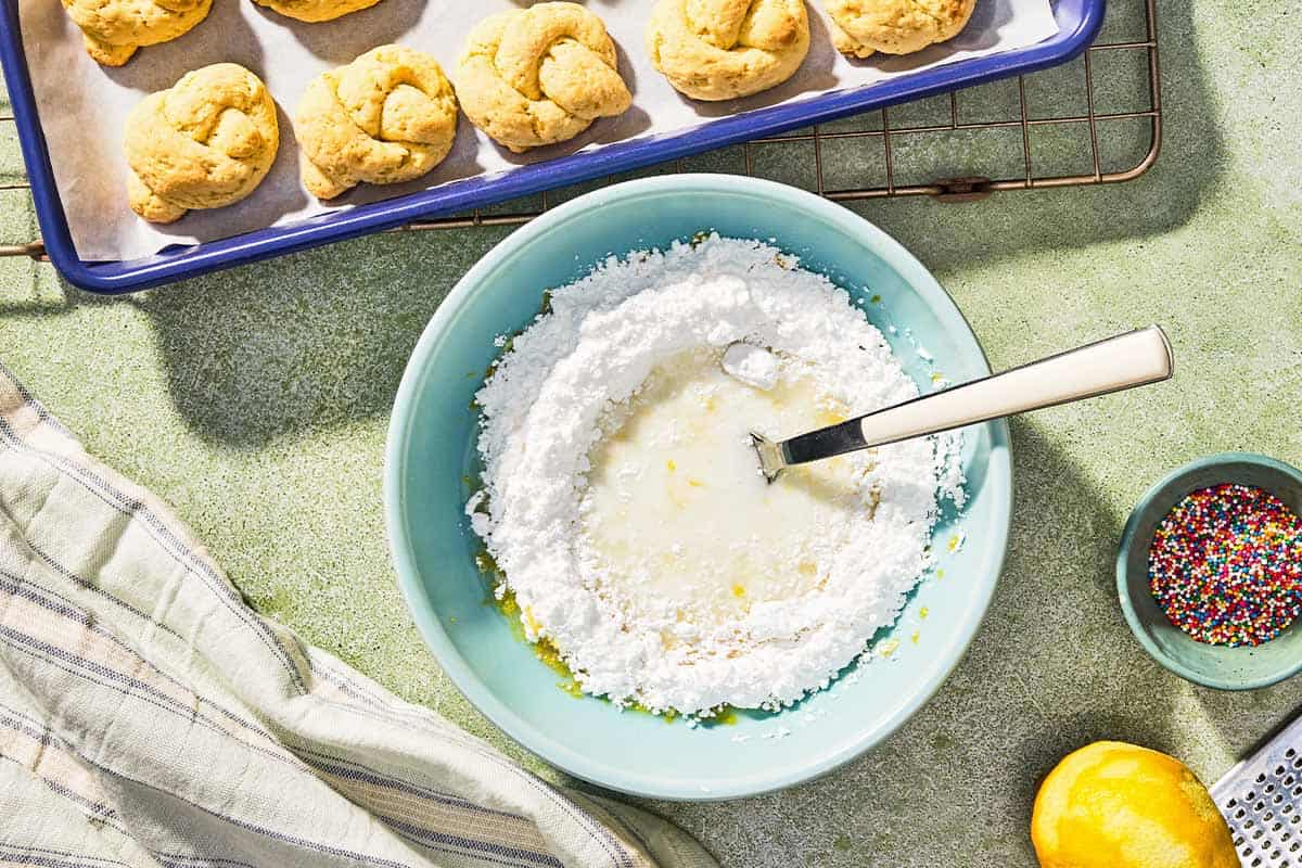 The icing for the Italian lemon cookies in a bowl with a spoon just before being mixed together. Next to this is a kitchen towel, a zested lemon, a bowl of non-pareils, and the undecorated cookies on a parchment lined sheet pan.