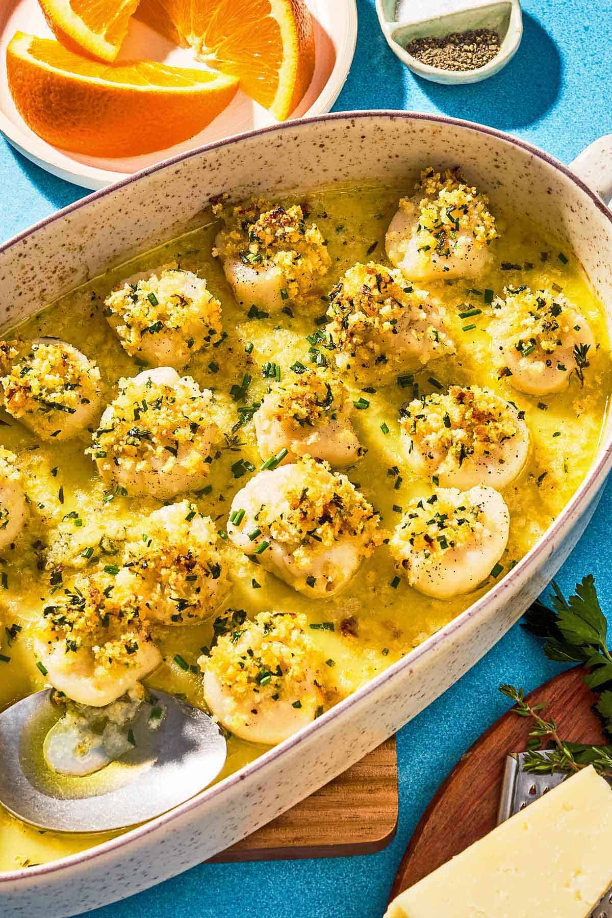 Baked scallops scented with orange and blanketed in fresh herbs, bread crumbs and Romano cheese rest in a white baking dish with a serving spoon. Next to this is a plate of orange wedges, a bowl of salt and black pepper, and a cutting board with a wedge of Pecorino Romano cheese and a cheese grater.