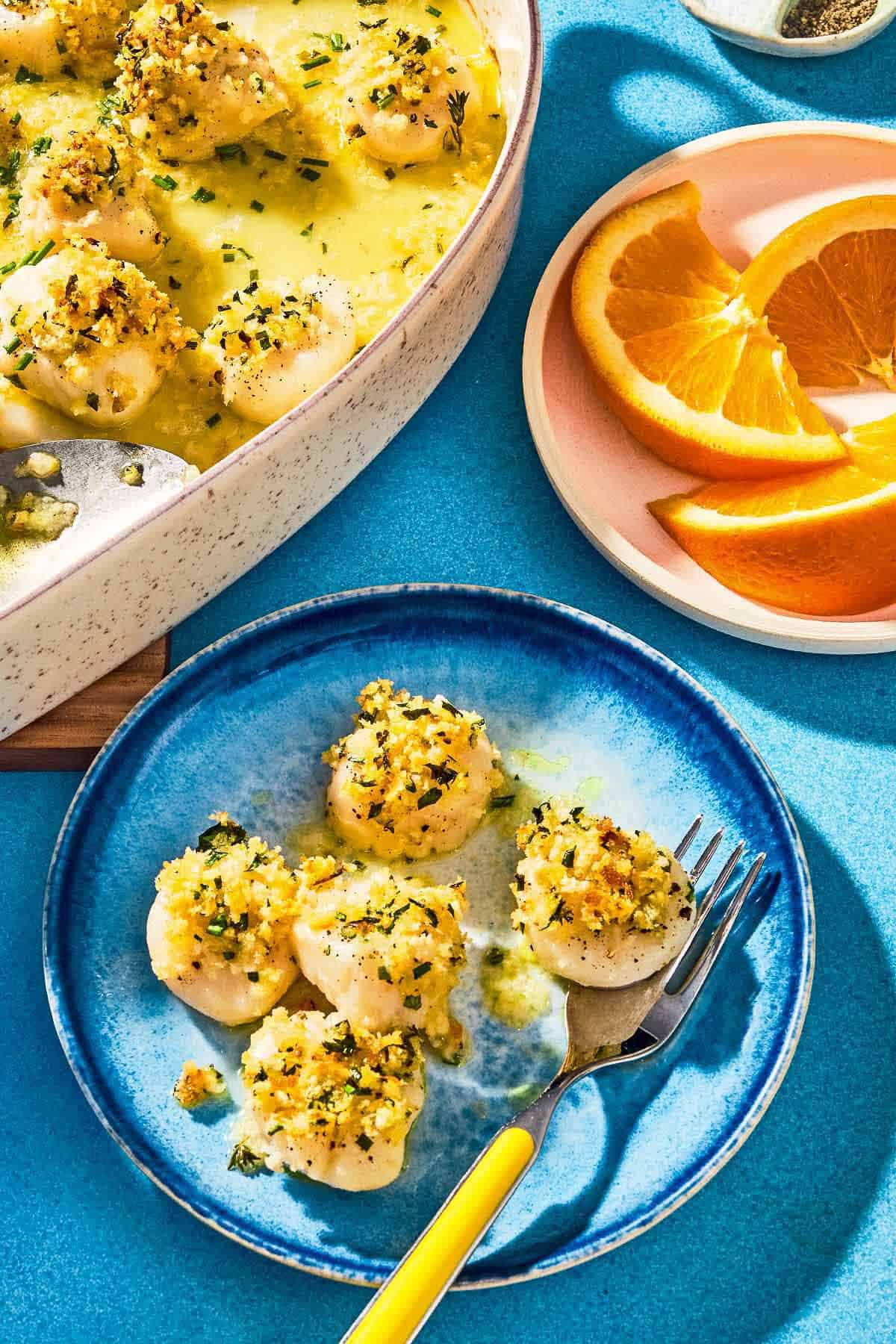 Five tender baked scallops on a plate with a fork. Next to this are the rest of the baked scallops in a baking dish, and a plate of orange wedges.
