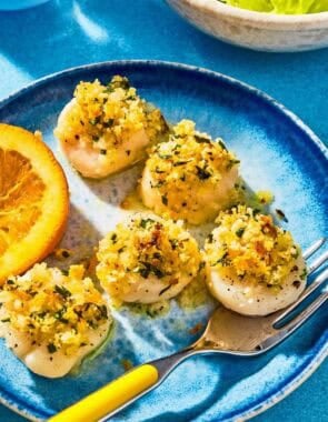 Five baked scallops on a plate with a fork and a lemon wedge.