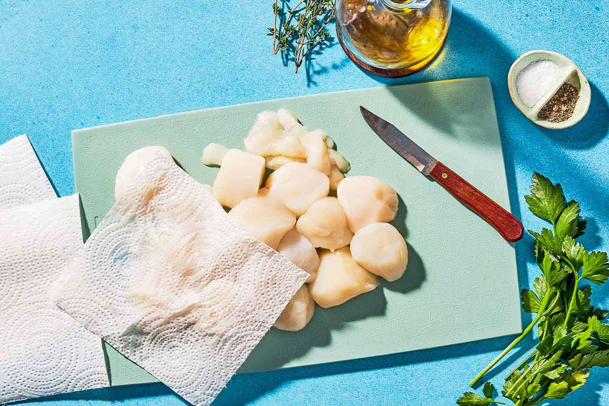 Uncooked scallops, paper towels and a knife on a cutting board next to sprigs of parsley, a bowl of salt and black pepper and a bottle of olive oil.