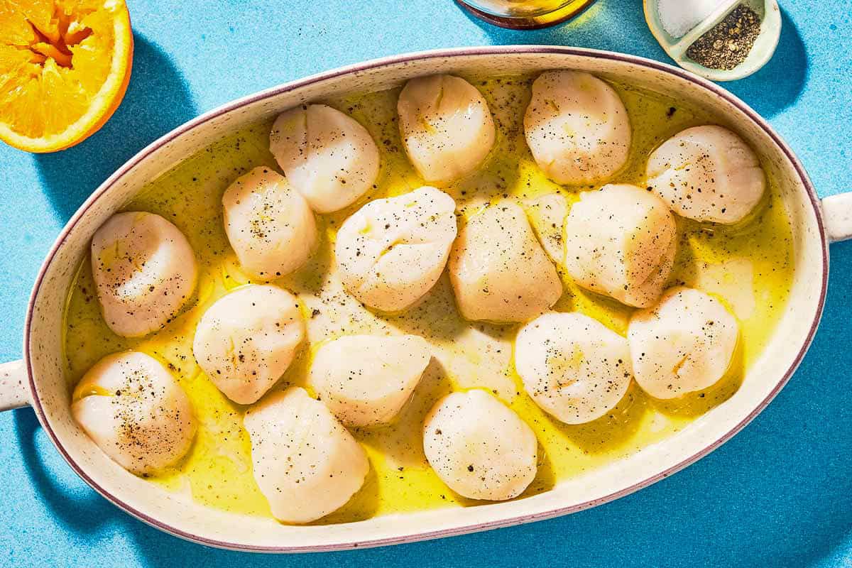 The uncooked scallops in a baking dish next to a juiced orange half and a bowl of salt and black pepper.