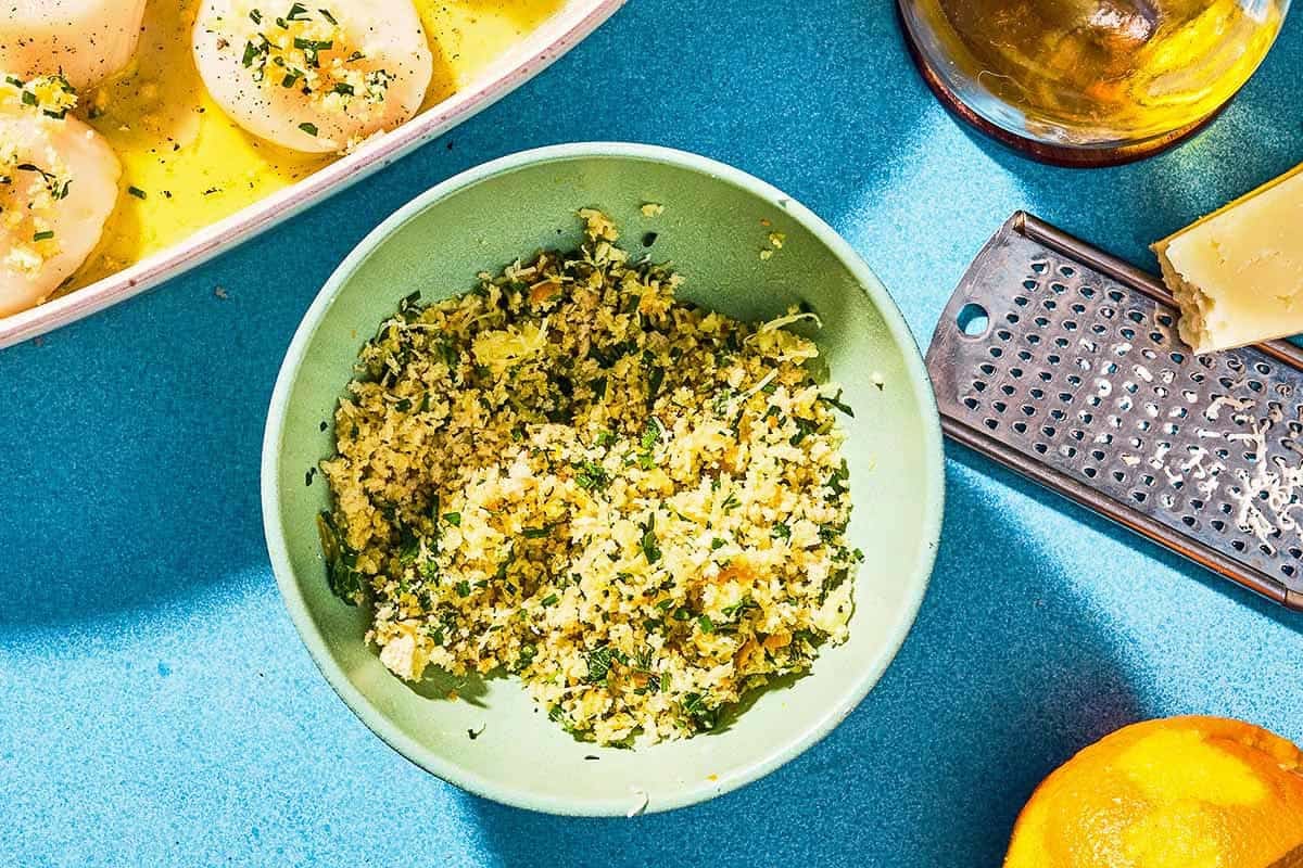 The breadcrumb topping for the baked scallops in a bowl next to an orange, Pecorino Romano cheese with a cheese grater, a bottle of olive oil, and the uncooked scallops in a baking dish.