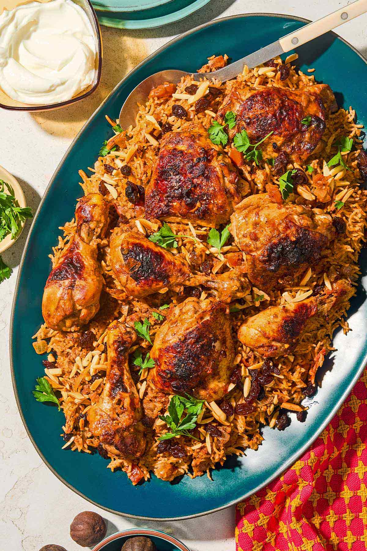Kabsa, a chicken and rice dish knowns as the National dish of Saudi Arabia, is commonly found throughout Middle Eastern countries and the Levant. In this photo, kabsa is piled on a deep blue platter and topped with parsley. A serving spoon is tucked into the side, and a bowl of yogurt, commonly served alongside Kabsa recipes is in the upper corner of the image.