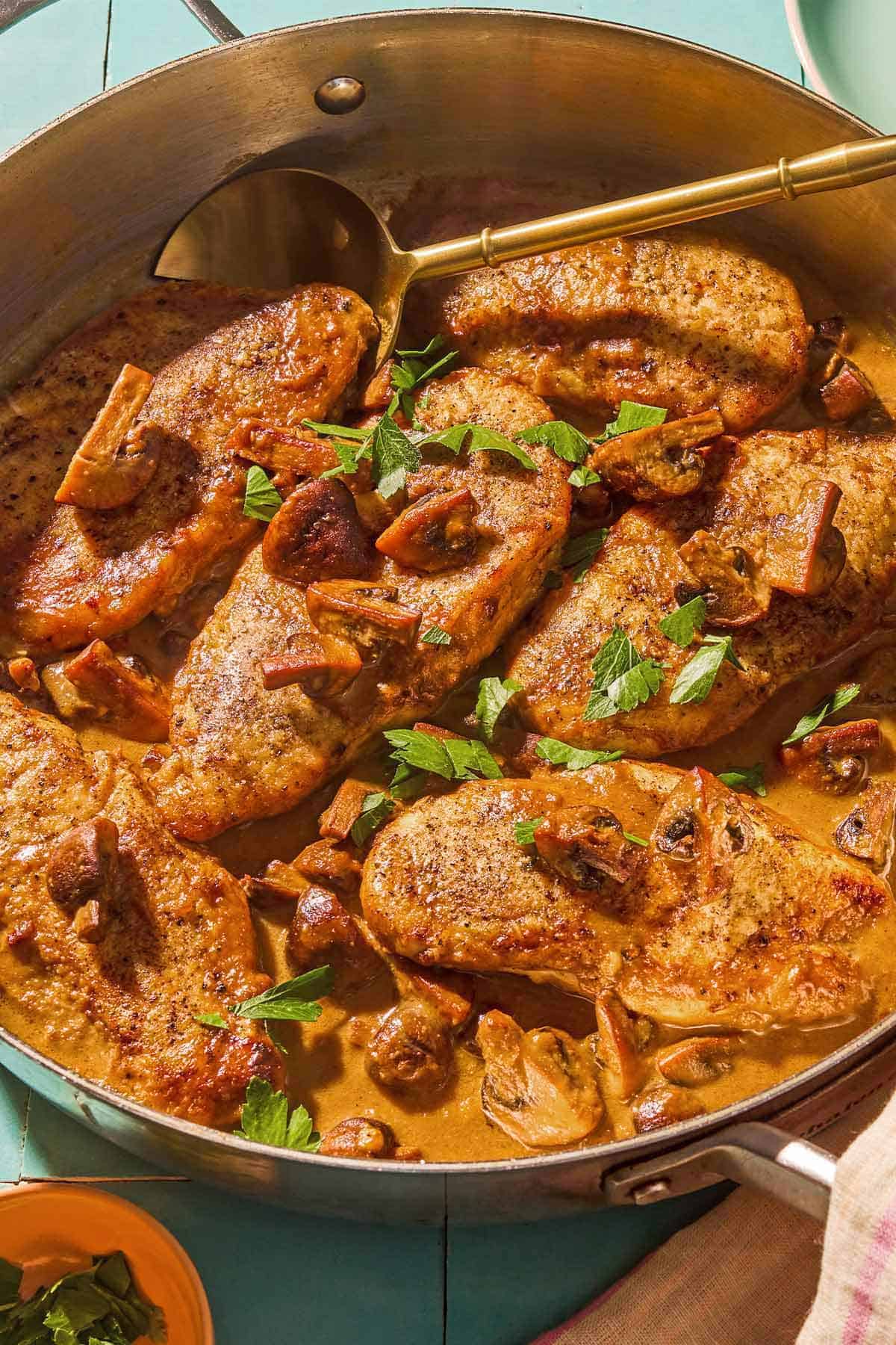 Classic chicken marsala in a skillet with a gold serving serving spoon. Quartered mushrooms and minced parsley are spooned over the top of the chicken resting in a marsala wine sauce.