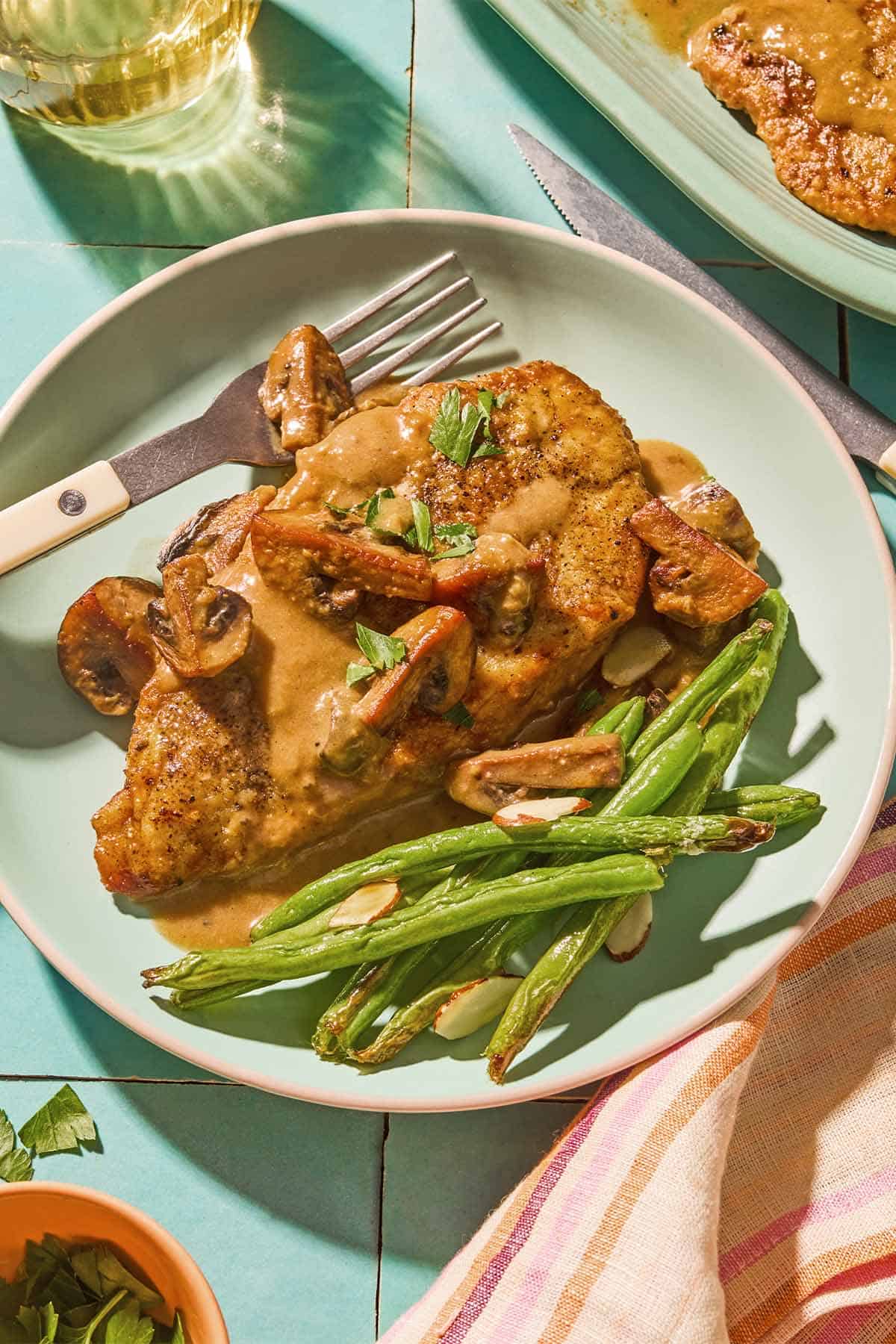 Chicken marsala on a plate with green beans and a fork. Next to this is a knife, a cloth napkin, a bowl of chopped parsley, a glass of wine, and the rest of the chicken marsala on a platter.