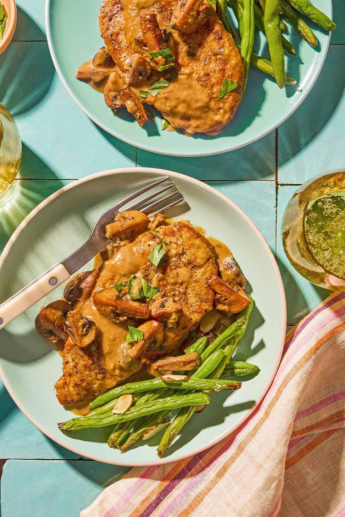 Two servings of chicken marsala on plates with green beans. One also has a fork.