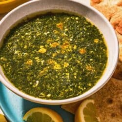 A bowl of molokhia in a bowl on a plate with pita and lemon wedges.