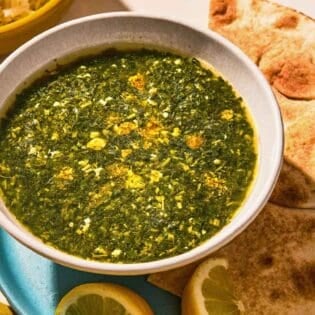 A bowl of molokhia in a bowl on a plate with pita and lemon wedges.