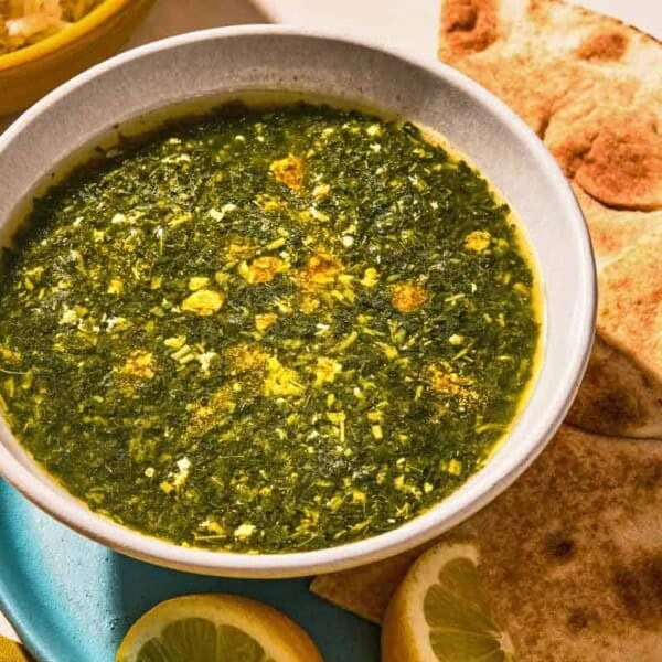 A bowl of molokhia in a bowl on a plate with pita and lemon wedges.