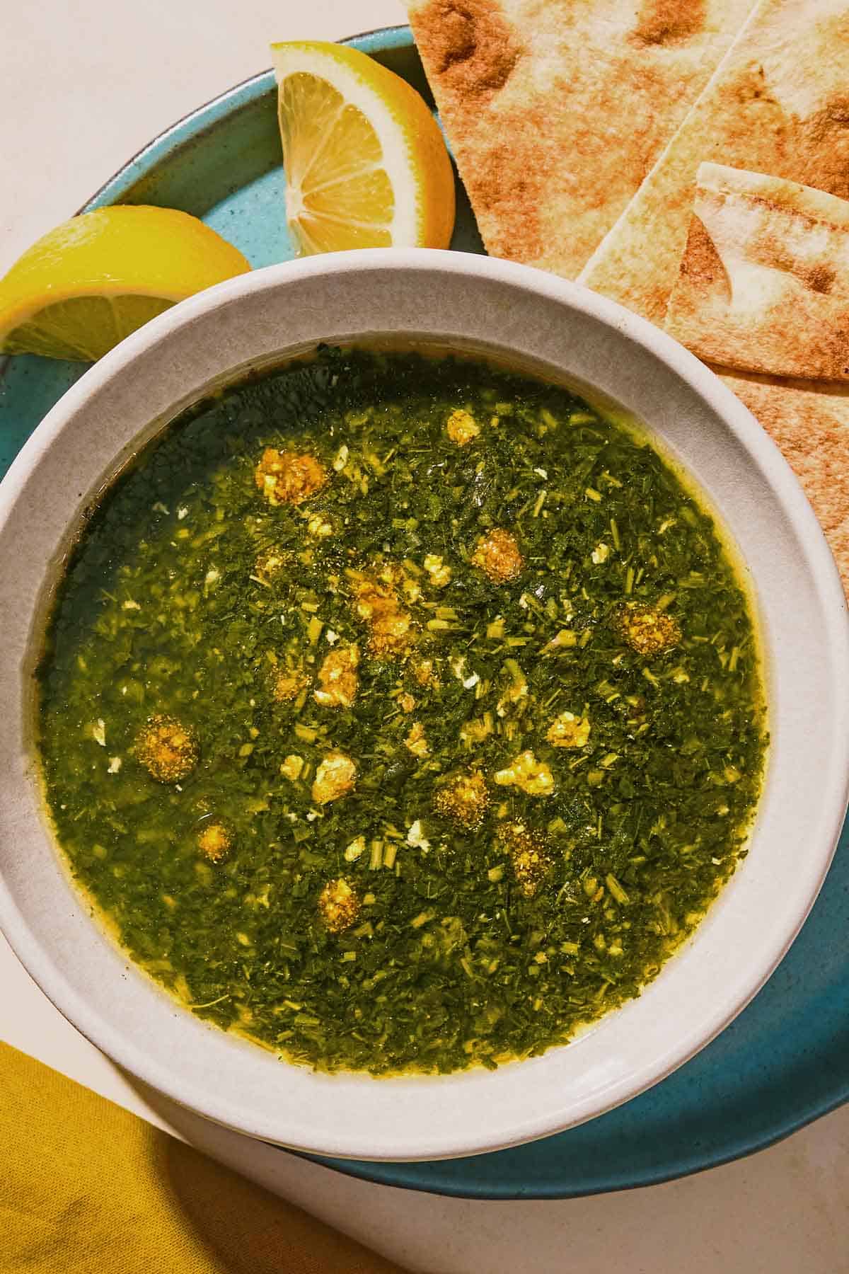 A bowl of molokhia in a bowl on a plate with pita and lemon wedges.