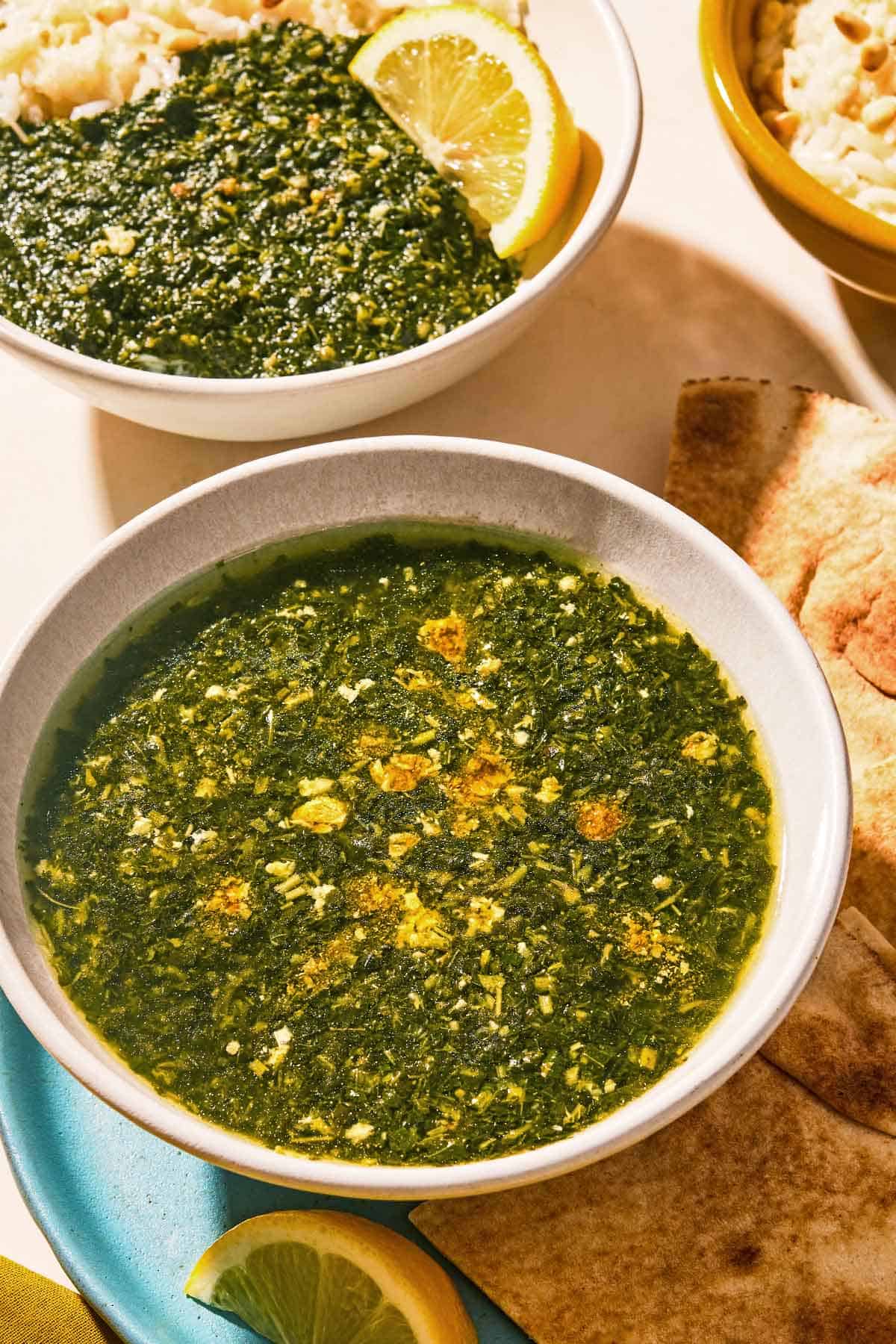 A bowl of molokhia in a bowl on a plate with pita and lemon wedges. Next to this is another bowl of the molokhia with rice and a lemon wedge, and a bowl of the rice.