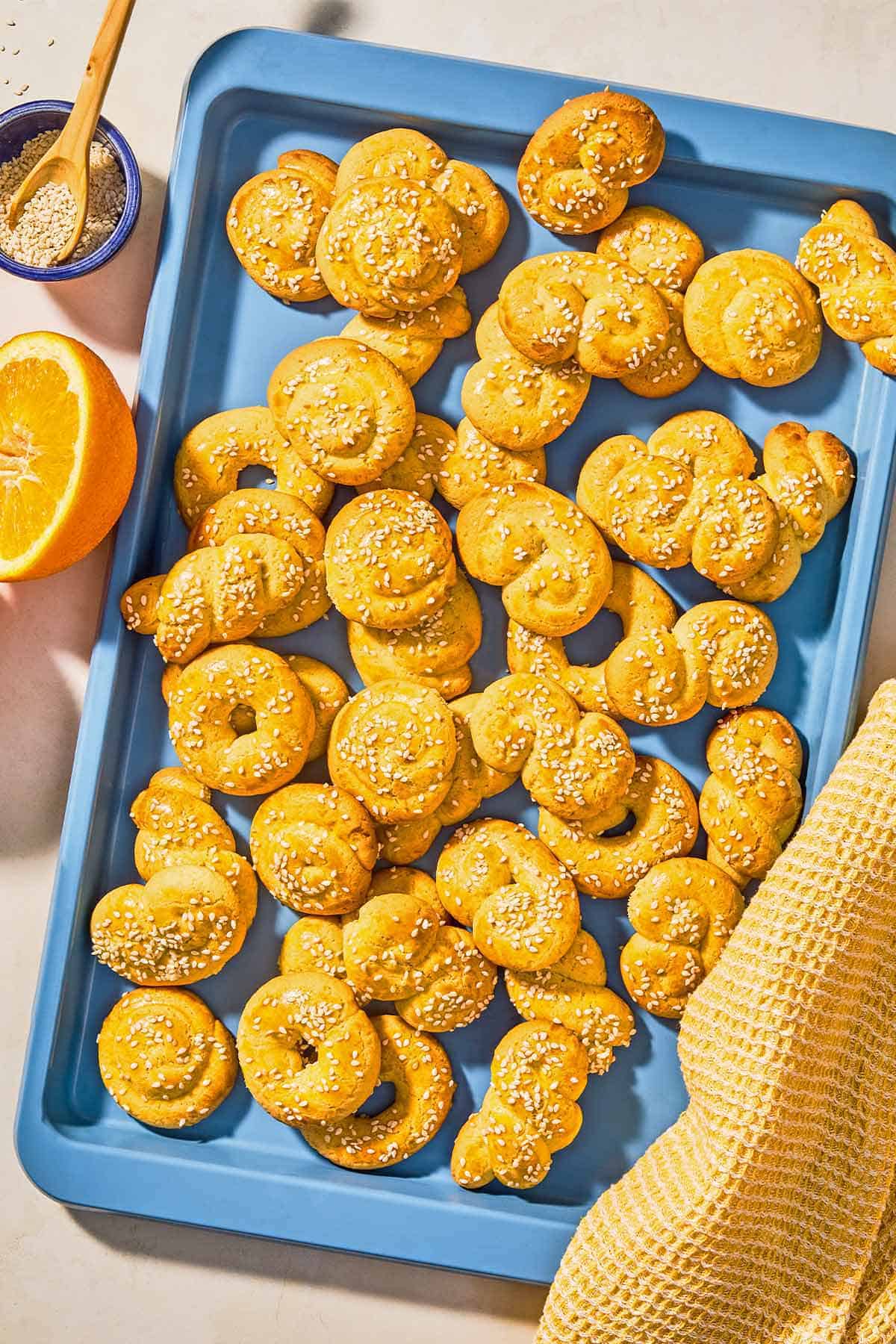 Baked Koulourakia topped with sesame seeds a sheet pant next to an orange half and a bowl of sesame seeds with a spoon.