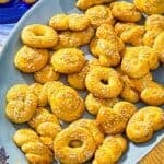 Koulourakia on a platter next to a plate with 2 koulourakia and a bowl of sesame seeds with a spoon.