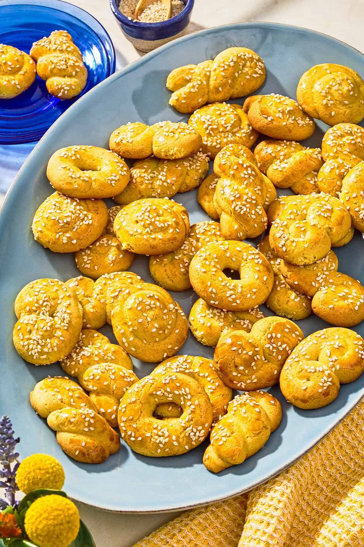 Koulourakia on a platter next to a plate with 2 koulourakia and a bowl of sesame seeds with a spoon.