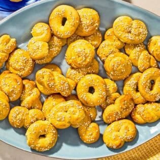 Koulourakia on a platter next to a bowl of sesame seeds with a spoon.
