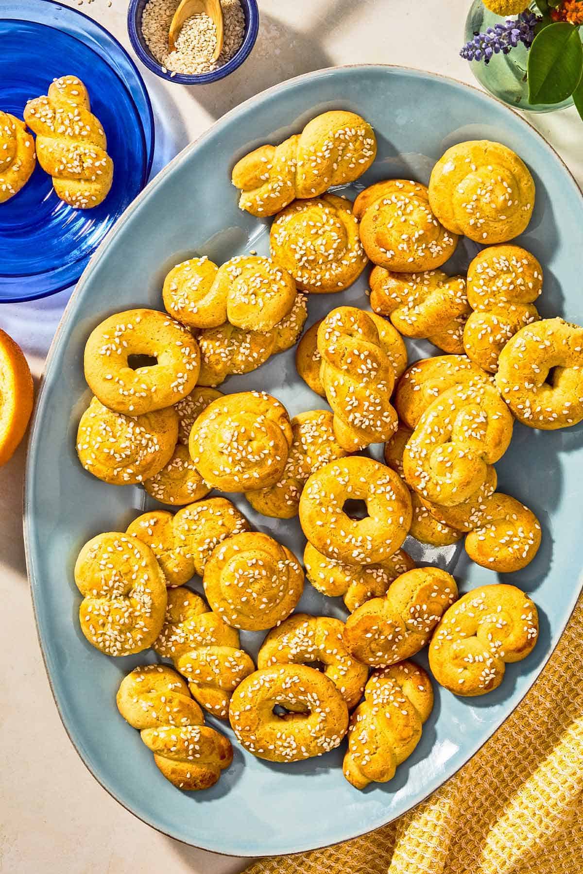 Koulourakia on a platter next to a plate of koulourakia and a bowl of sesame seeds with a spoon.