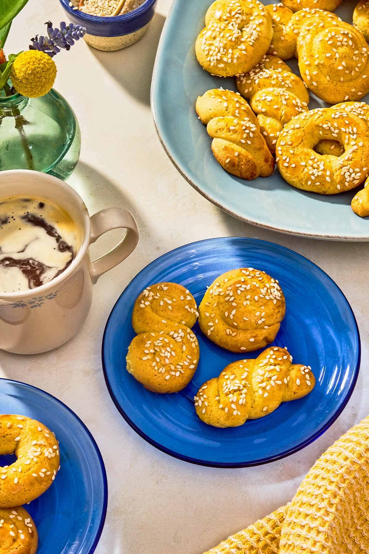 Three Koulourakia topped with sesame seeds on a plate. Next to this is a cup of coffee and a platter with the rest of the koulourakia.