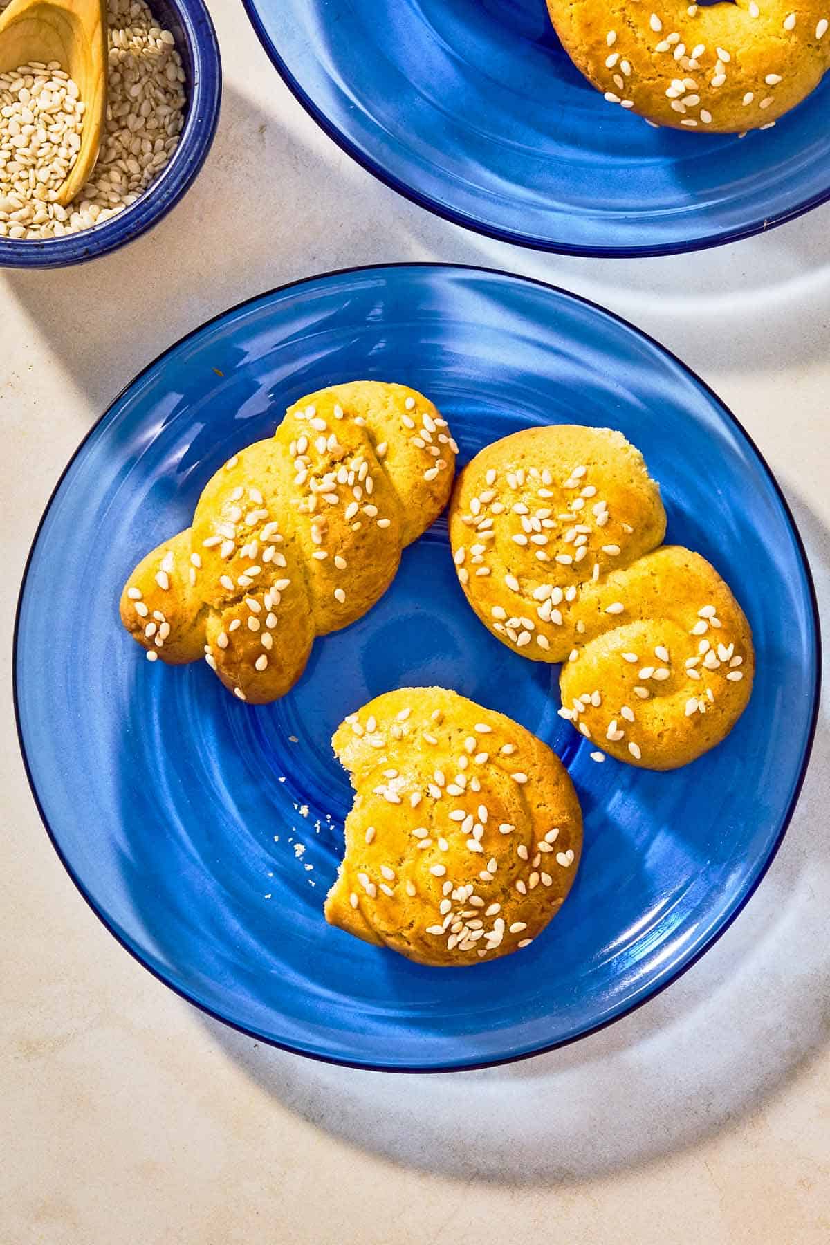 Three Koulourakia topped with sesame seeds on a plate next to another plate of the koulourakia and a bowl of sesame seeds with a spoon.