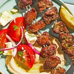 Lamb kabobs on a plate with a lemon wedge, tomato salad and hummus.