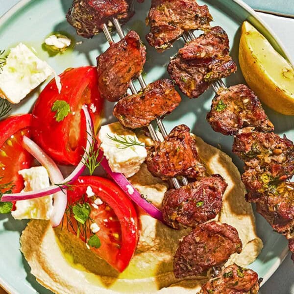 Lamb kabobs on a plate with a lemon wedge, tomato salad and hummus.
