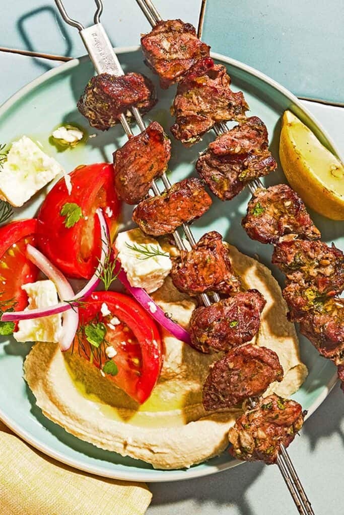 Lamb kabobs on a plate with a lemon wedge, tomato salad and hummus.