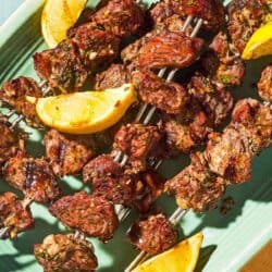 Several lamb kabobs on a platter with lemon wedges.