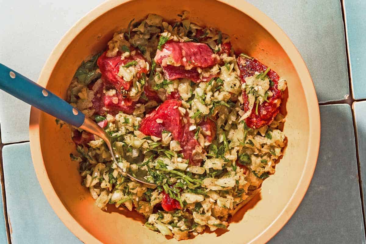 The uncooked cubed lamb being mixed together with the garlic spice rub in a bowl with a spoon.