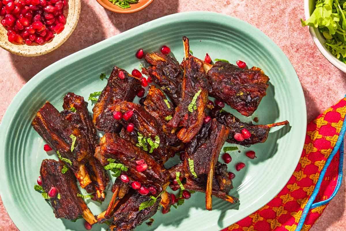 Lamb ribs on a platter next to a bowl of pomegranate ariels, mint and lettuce.