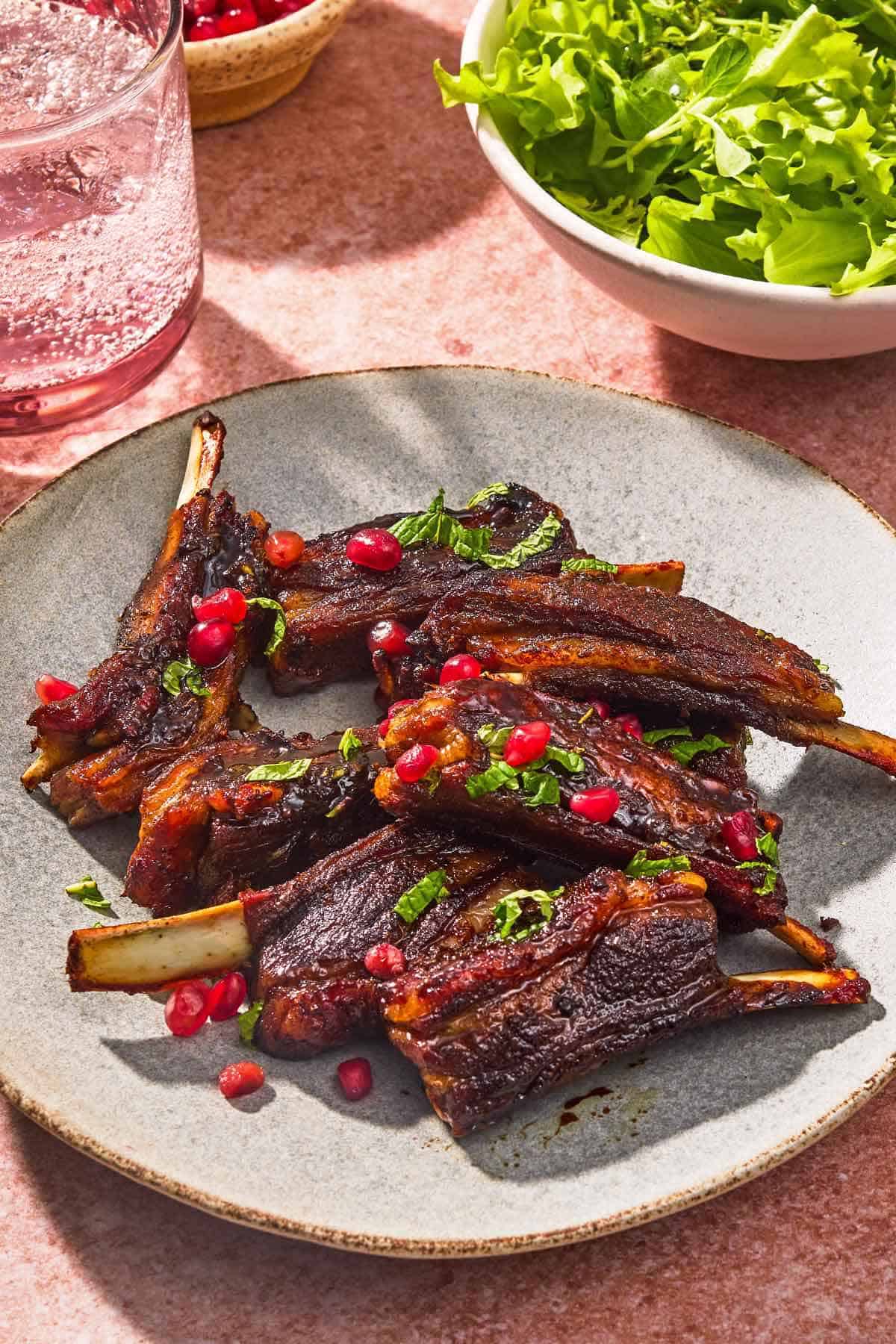A serving of lamb ribs on a plate next to a glass of sparkling water and a bowl of lettuce.