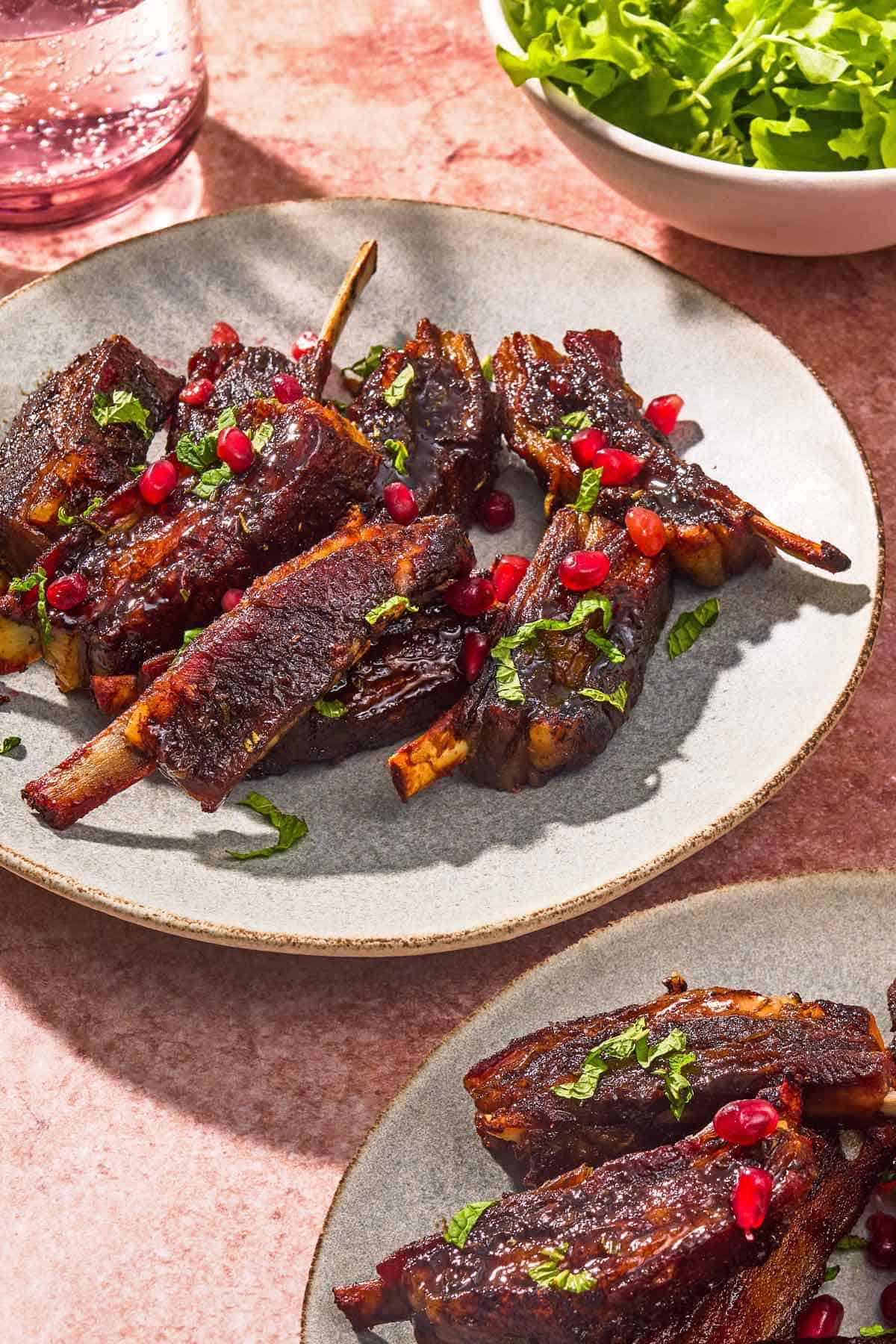 A serving of lamb ribs on a plate next to a glass of sparkling water, a bowl of lettuce, and another plate of the ribs.