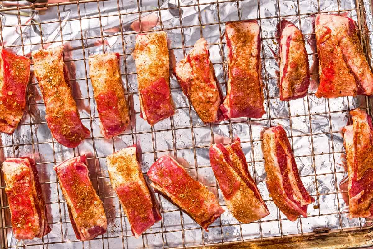 The seasoned, uncooked lamb ribs on a wire rack over a foil lined sheet pan.