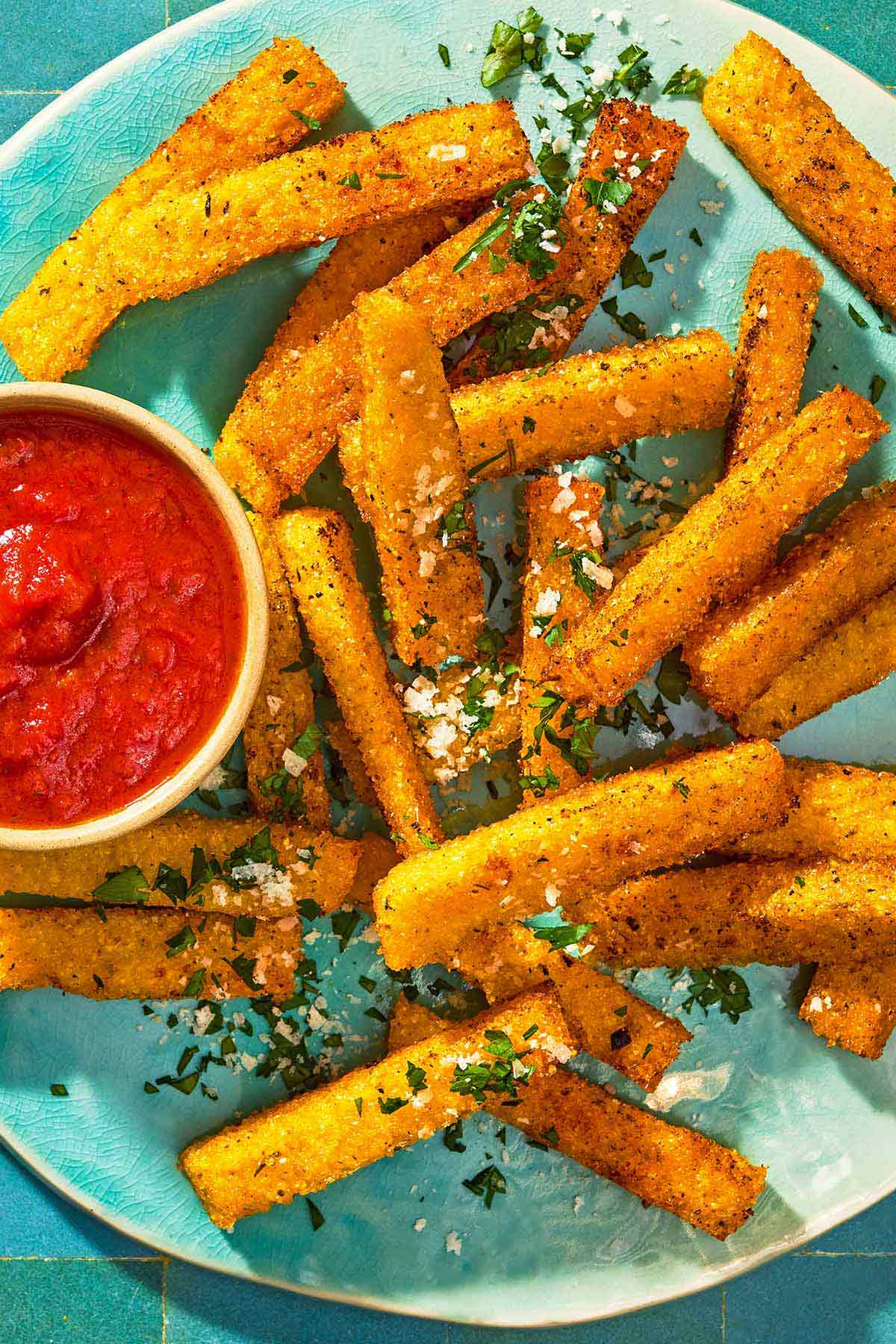 Polenta fries topped with grated parmesan cheese and parsley on a platter with a bowl of marinara sauce.