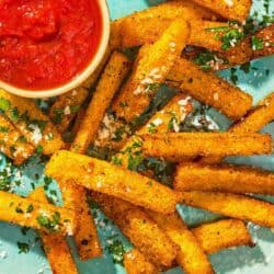 Polenta fries topped with grated parmesan cheese and parsley on a platter with a bowl of marinara sauce.