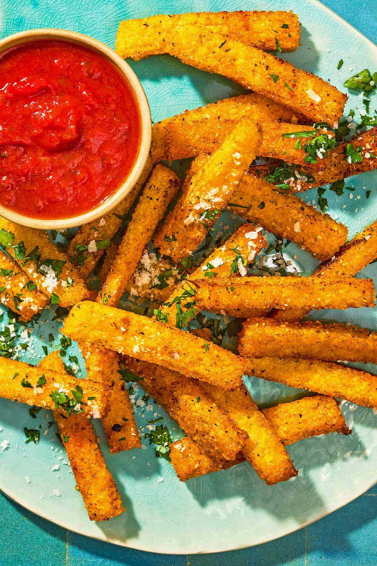 Polenta fries topped with grated parmesan cheese and parsley on a platter with a bowl of marinara sauce.
