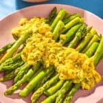 Sauce gribiche over asparagus on a plate next to a bowl of the sauce with a spoon and a bowl of salt and pepper.