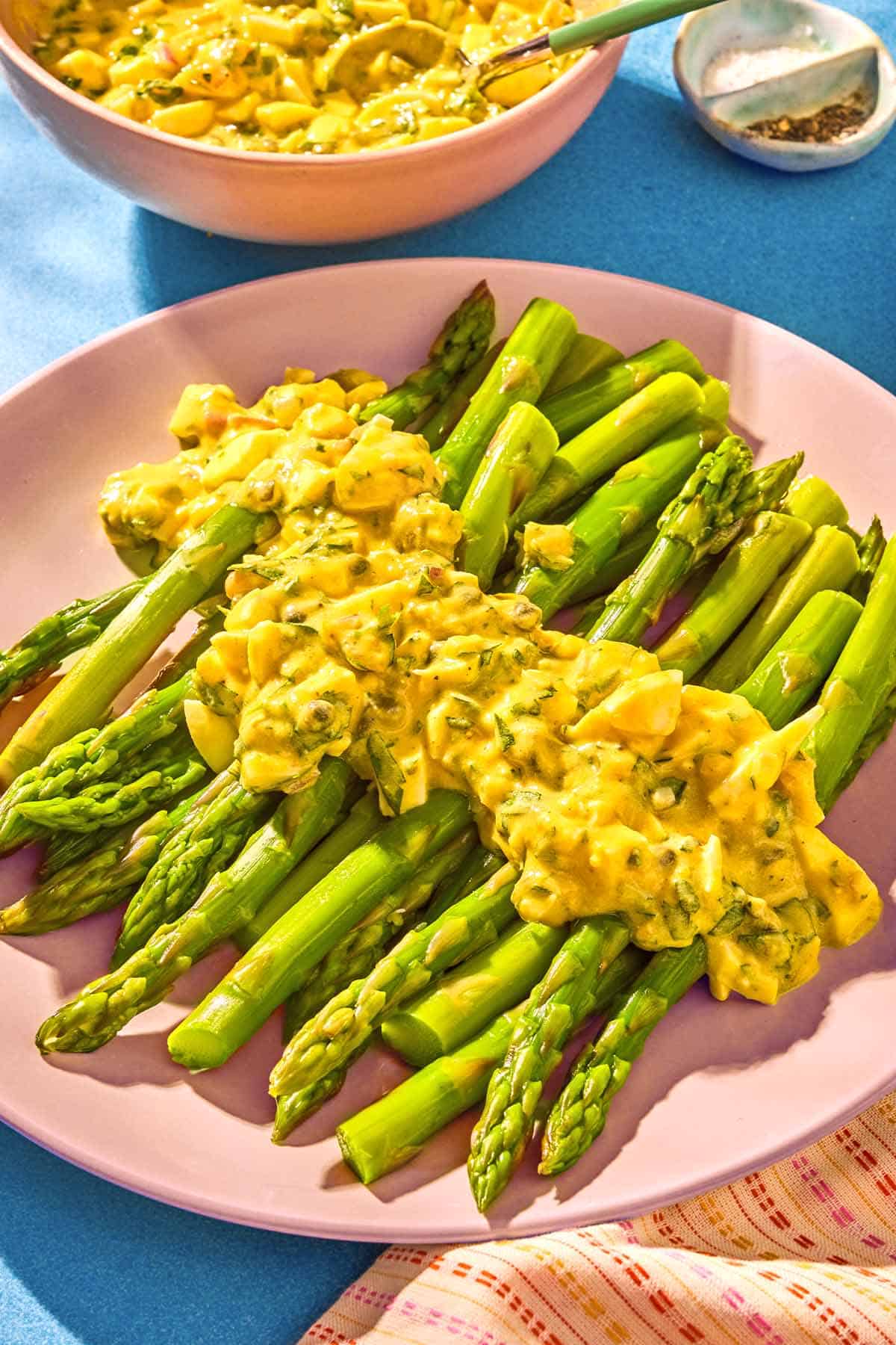 Sauce gribiche over asparagus on a plate next to a bowl of the sauce with a spoon and a bowl of salt and pepper.