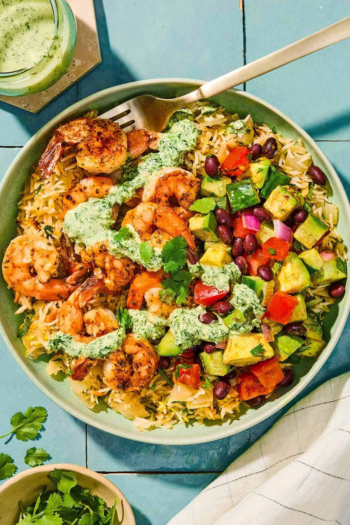 A shrimp bowl with a fork next to a jar of green goddess dressing with a spoon and a bowl of cilantro.