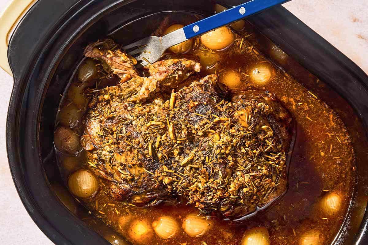The boneless leg of lamb recipe, pearl onions and broth mixture in a slow cooker with a fork.