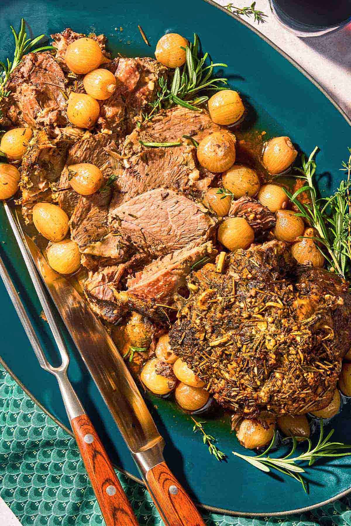 The sliced boneless leg of lamb with fresh herbs and pearl onions on a platter. with carving utensils.