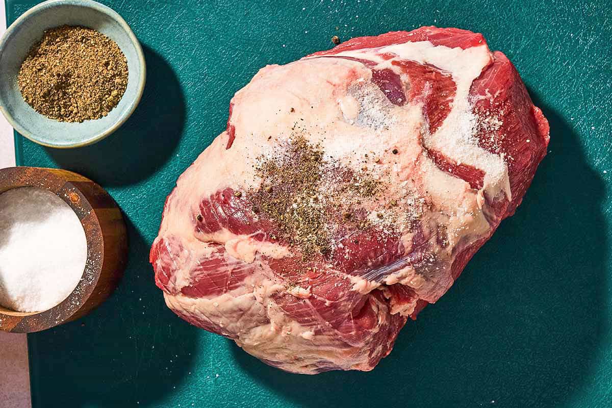 The uncooked boneless leg of lamb seasoned with salt and pepper on a cutting board with bowls of the salt and pepper.