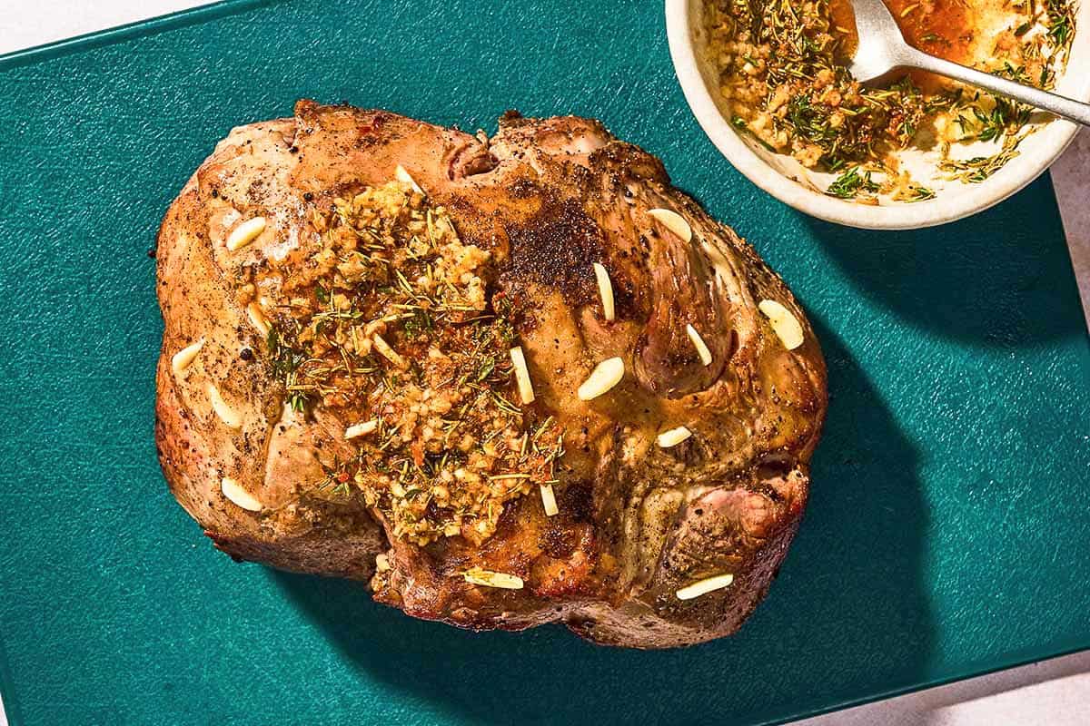 The seared boneless leg of lamb stuffed with sliced garlic and topped with the garlic spice rub on a cutting board with a bowl of the garlic spice rub.
