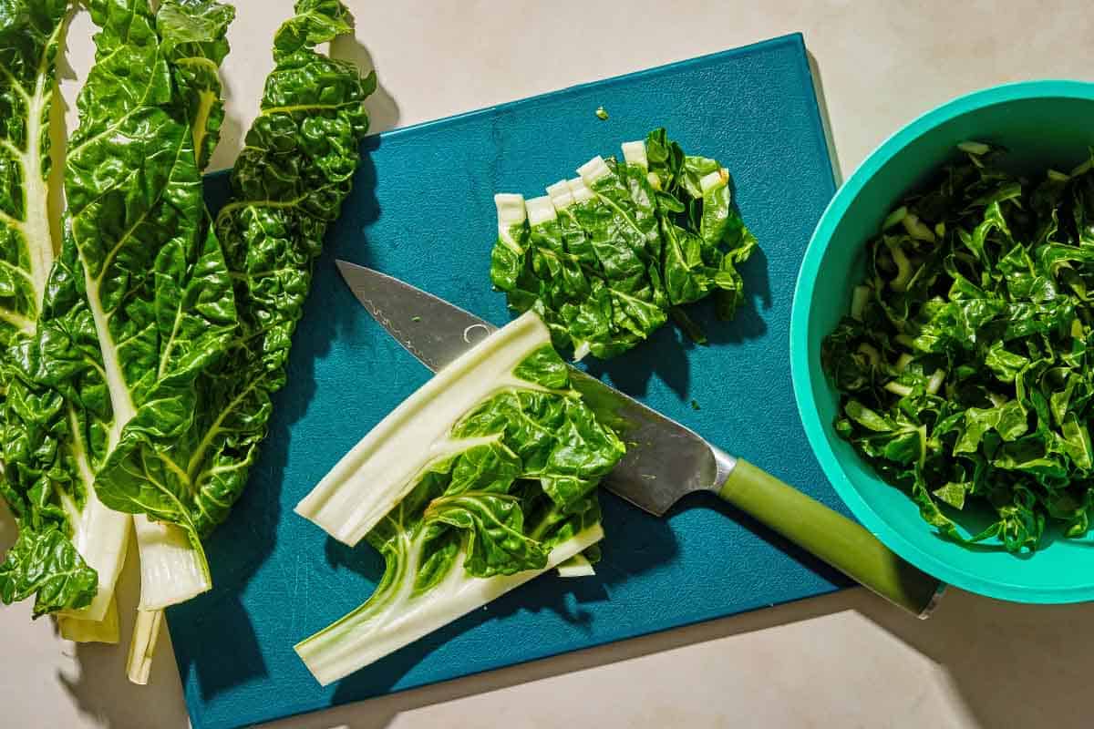 partially chopped stalks of chard on a cutting board with a knife. Next to this is some full chard stems, and a bowl of the chopped chard.